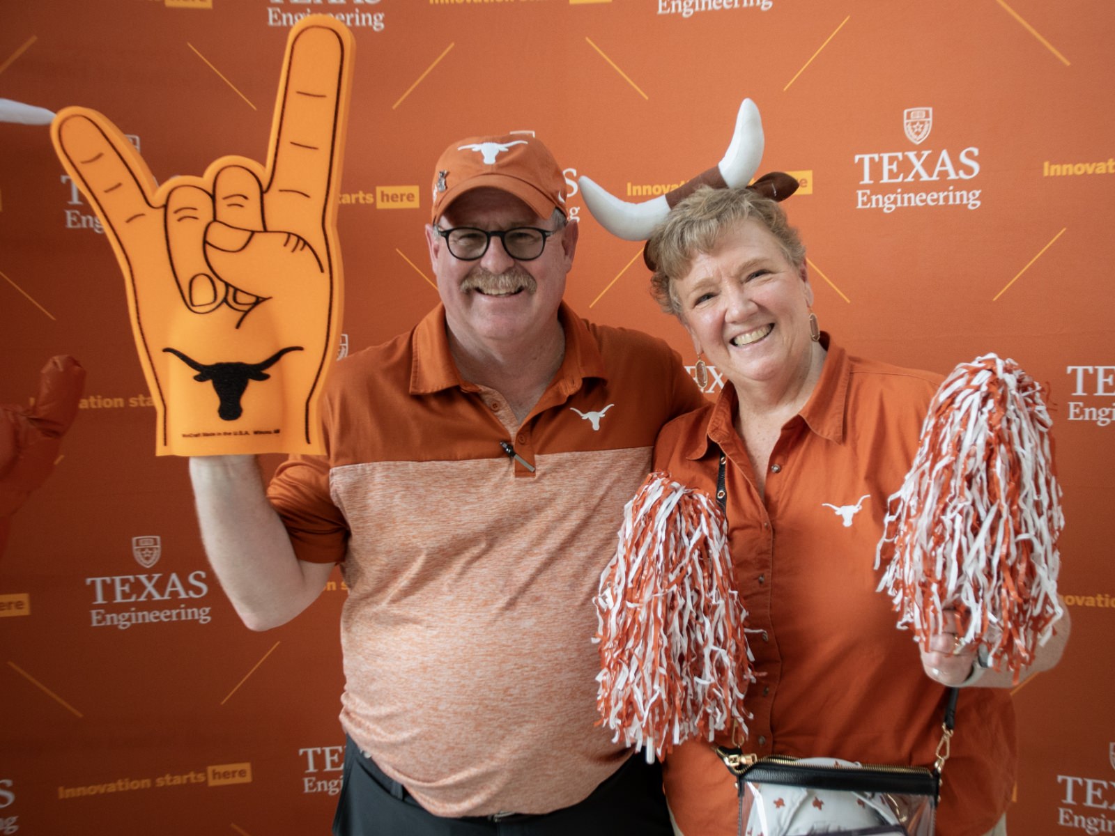 Texas engineering alumni at with UT Austin shirts smiling with pom-poms and a foam finger in from of Texas Engineering backdrop