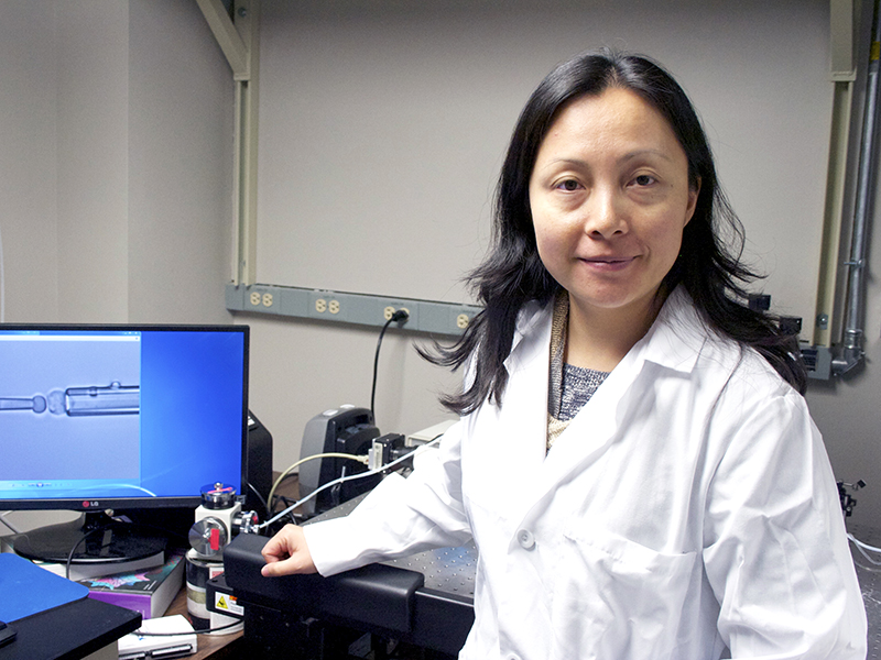 Portrait of Jiang Jenny in her lab in front of her computer while holding onto her desk.