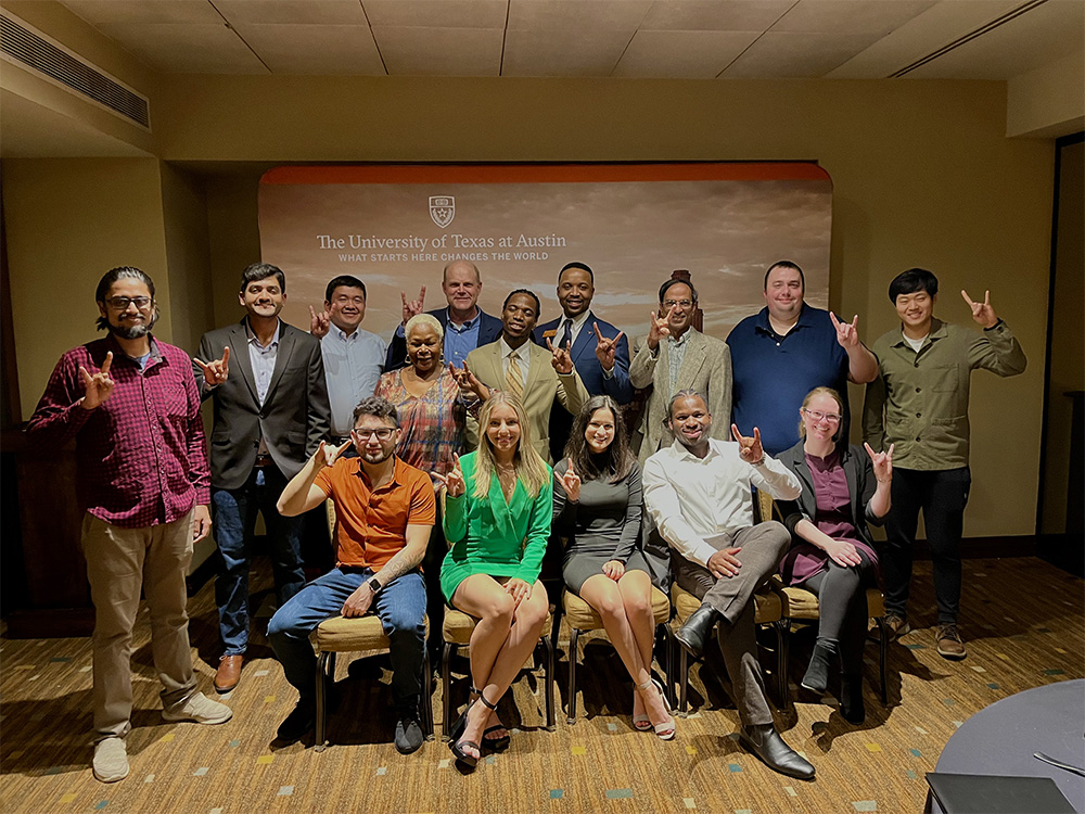 Group of people doing hook 'em horns hand sign in front The University of Texas at Austin sign