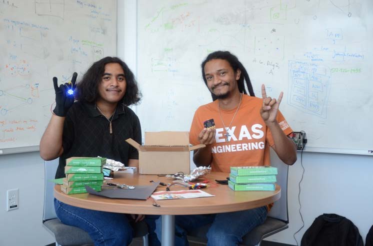 Two students hold their hands up in a hook em while work on an electrical device