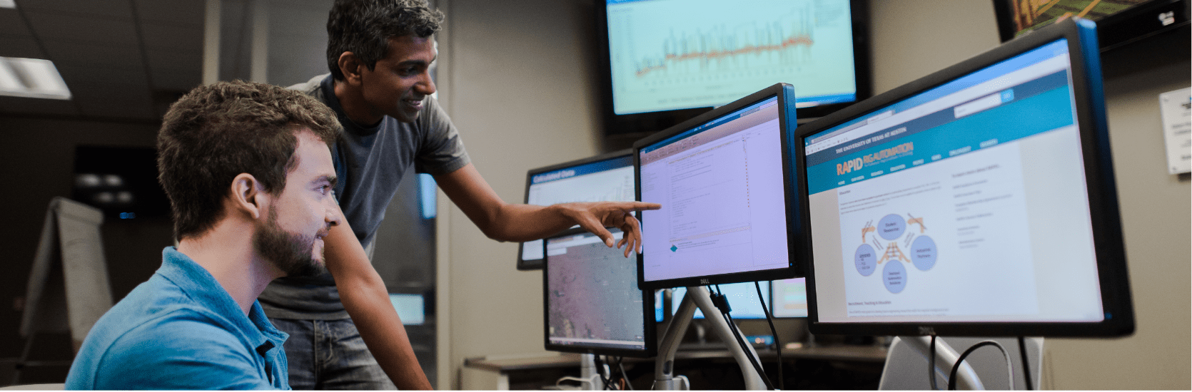 man stands and points at computer screen while other man looks on