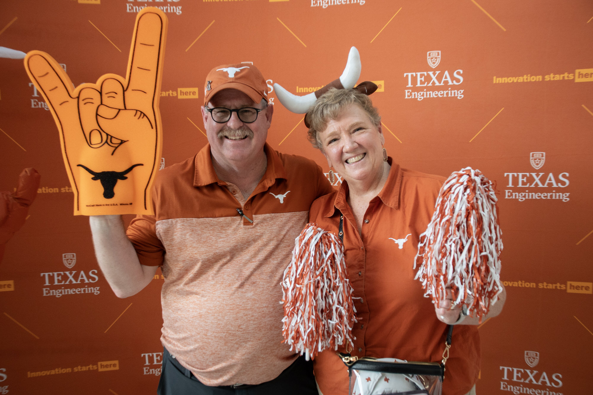 Texas engineering alumni at with UT Austin shirts smiling with pom-poms and a foam finger in from of Texas Engineering backdrop