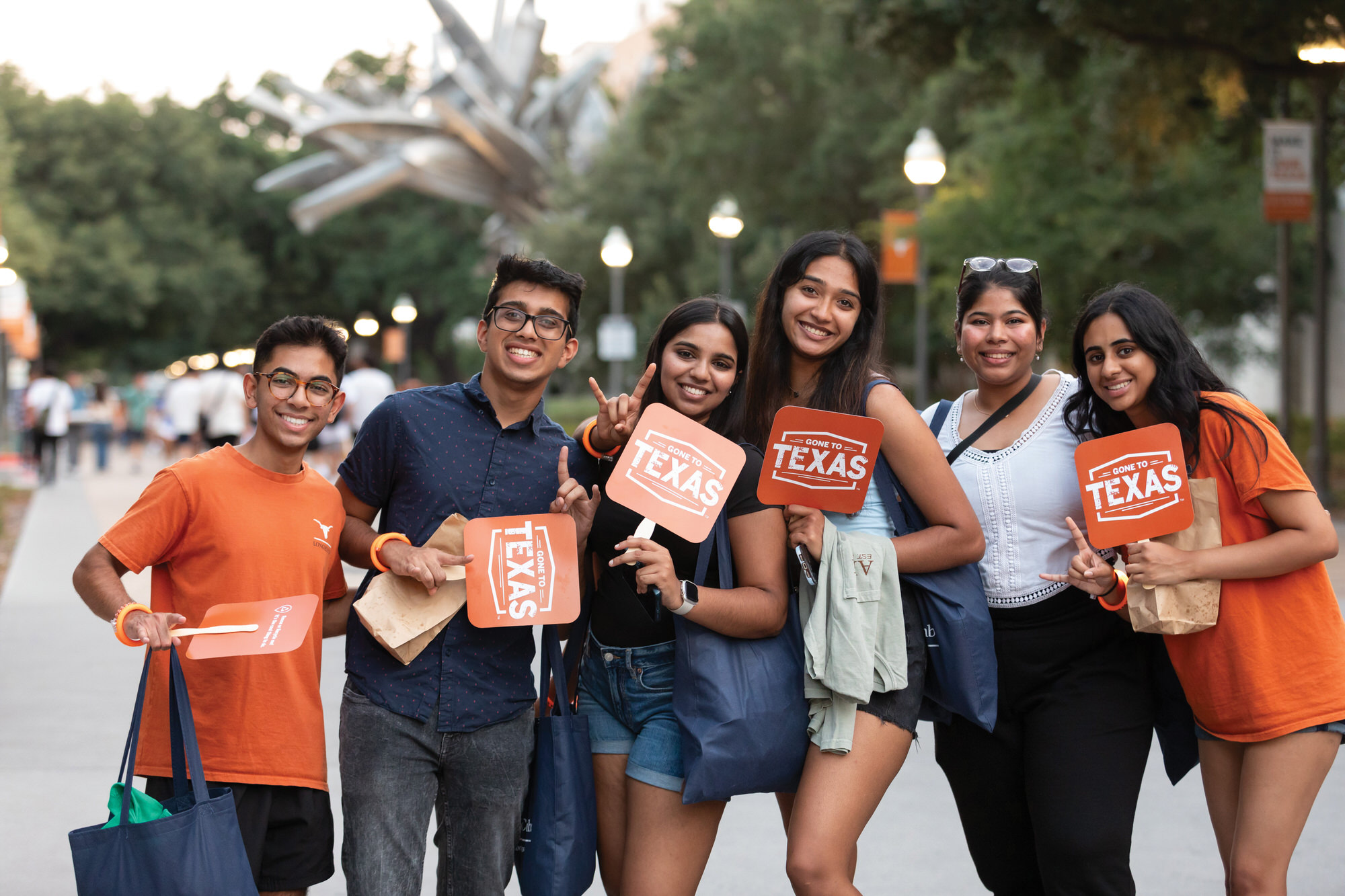 A diverse group of six UT Austin students smiling and holding “Gone to Texas” signs during an outdoor campus event, wearing burnt orange and Longhorn-themed attire.