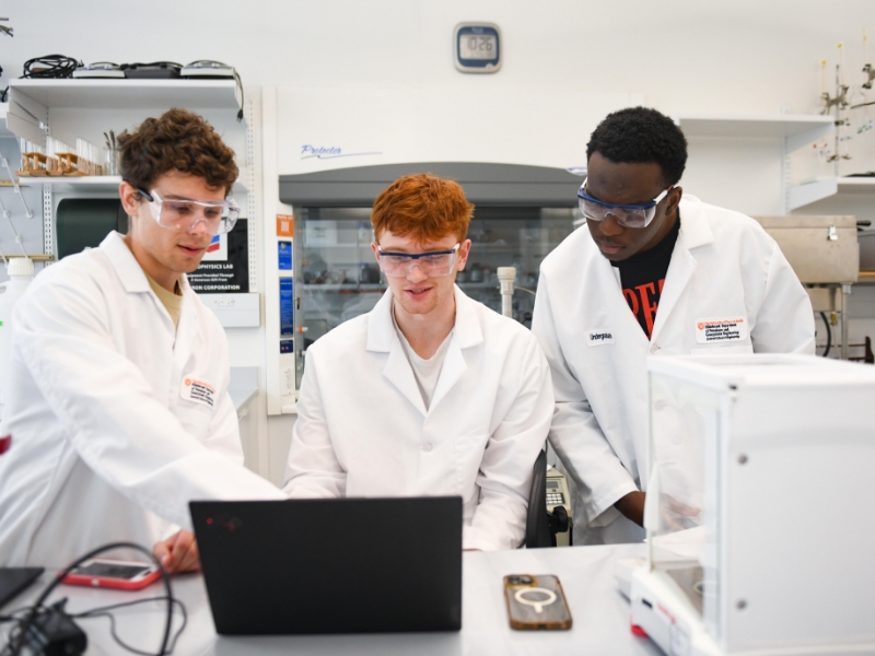 Texas Engineering students in a lab wearing safety goggles and lab coats gathered around a laptop