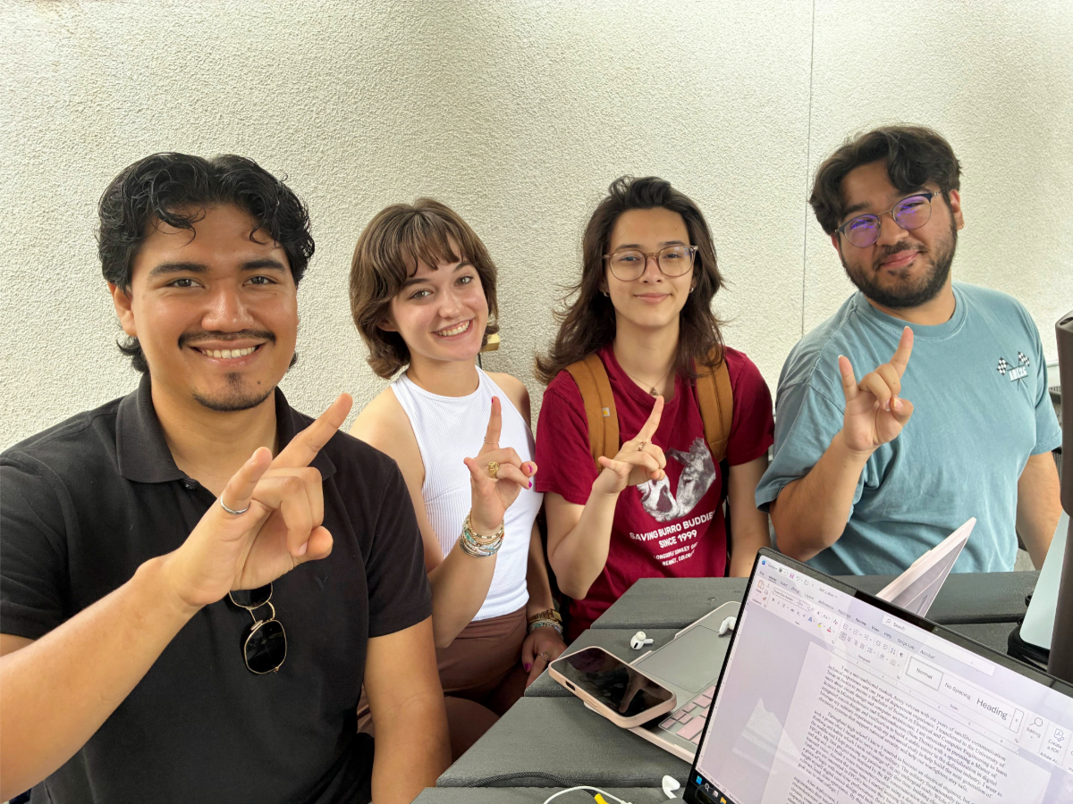 TAAP students sitting at table with laptops smiling and doing hook 'em horns hand signs. 