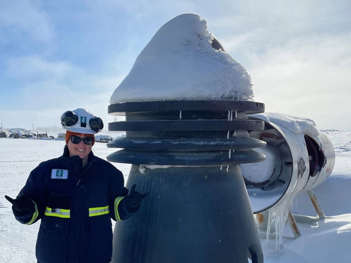 Texas Engineering alumna Jillian Jopling stands in a field of snow next to machinery used on the oil fields, in a hard hat and coat, throwing up two 
