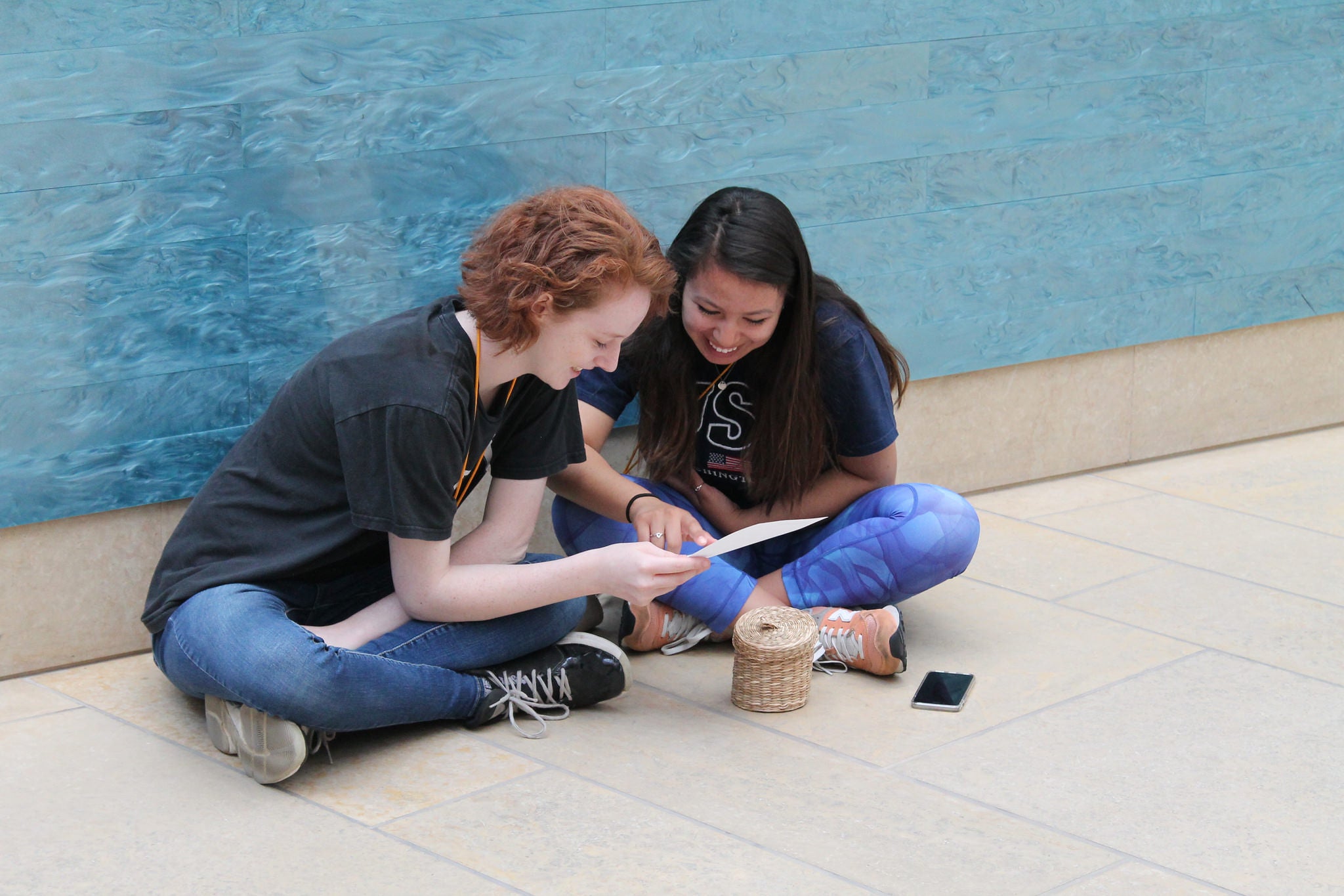 Two young students sitting on the floor looking at packet of papers together.